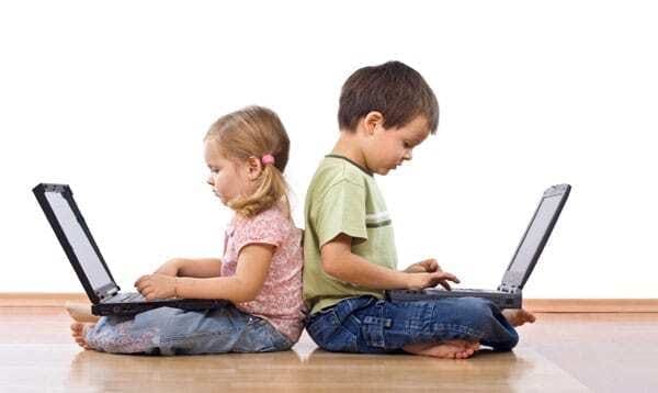 Duas crianças, uma menina e um menino, usando laptop. Crianças de costas umas para a outra, deixando de interagirem entre sim e interagindo pelo computador.